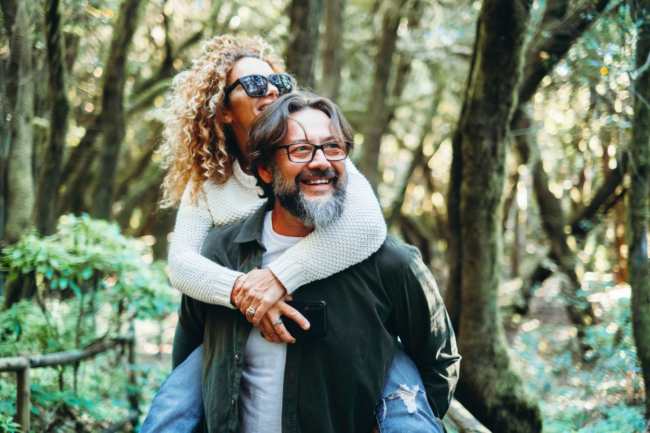 Smiling adult woman rides piggyback on bearded man in wooded setting.
