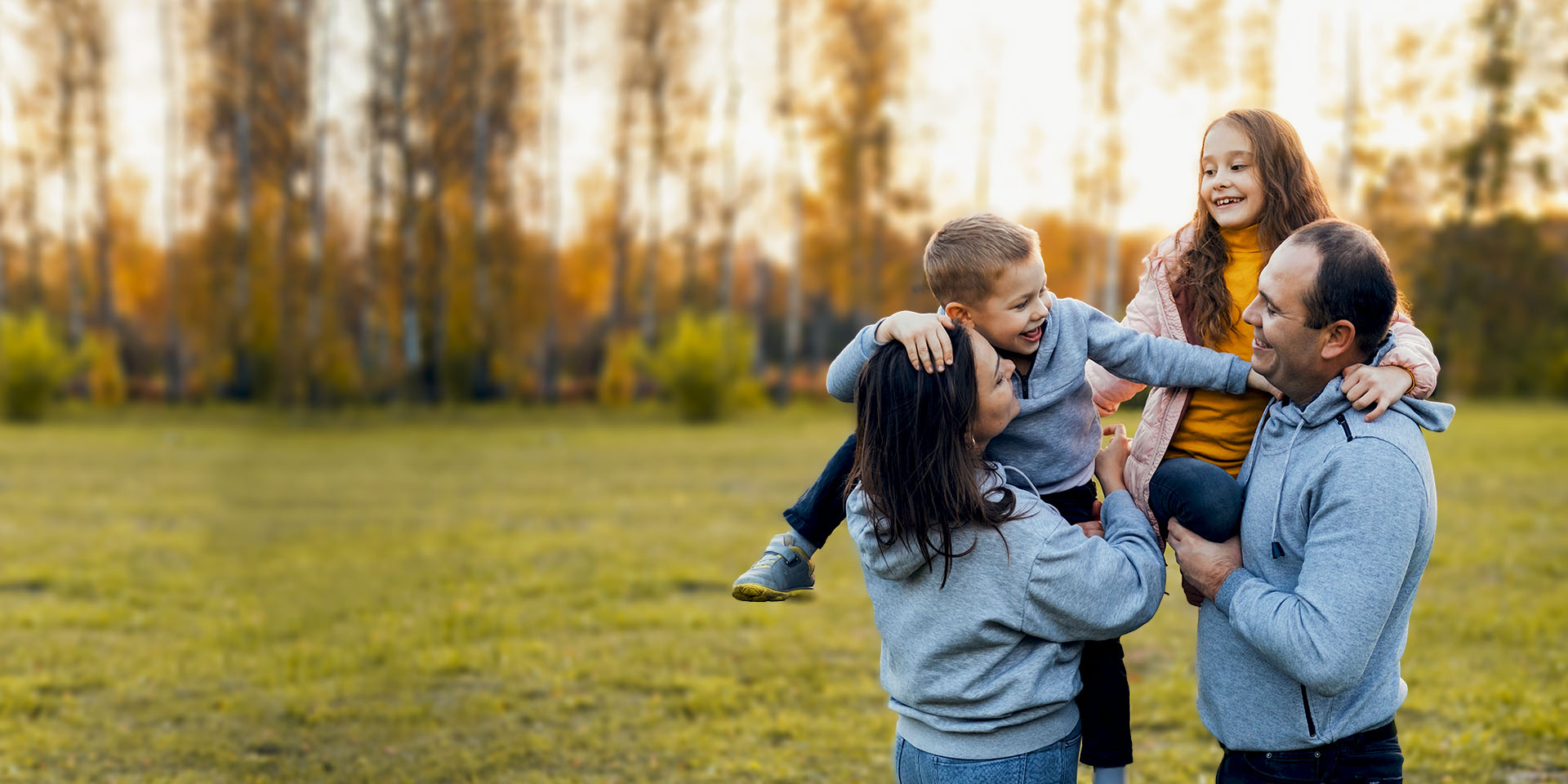 A mom, dad, daughter, and son stand in a field infront of a treeline on a fall day. The parents hold the kids; all smile.